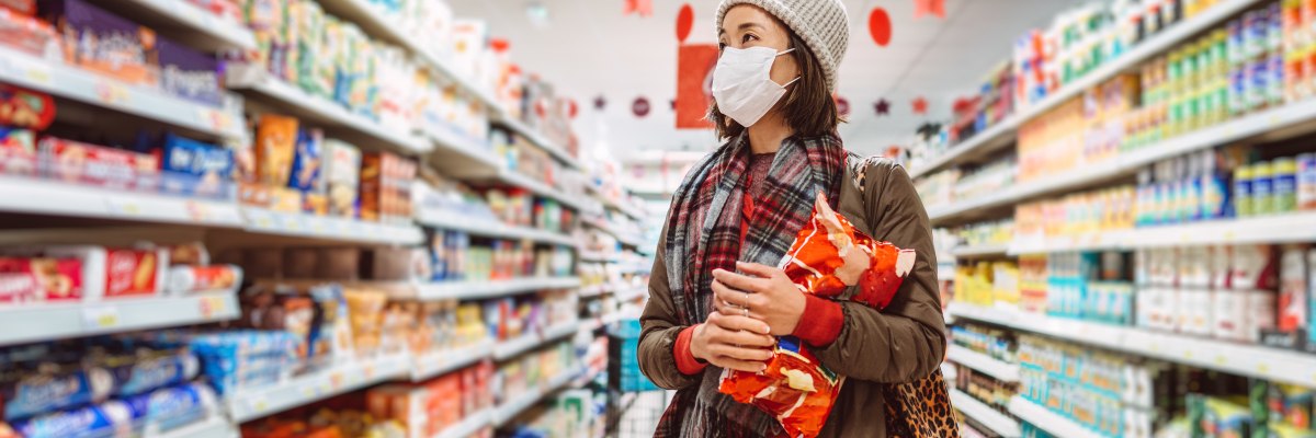 a lady with a mask and a hat on, holding a bag of crisps whilst standing in the packaged foods aisle in a hypermarket