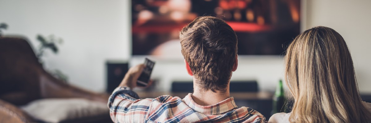 An image of a short haired man and long haired woman sat on a sofa watching something on a television as well as the man viewing something on his phone.